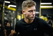 A focused man with short hair wearing black sportswear and wireless earphones stands in a gym. The background features gym equipment and blurred figures, with yellow and black tones dominating the setting.
