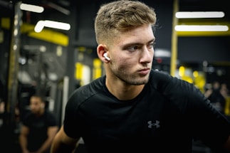A focused man with short hair wearing black sportswear and wireless earphones stands in a gym. The background features gym equipment and blurred figures, with yellow and black tones dominating the setting.