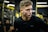 A focused man with short hair wearing black sportswear and wireless earphones stands in a gym. The background features gym equipment and blurred figures, with yellow and black tones dominating the setting.