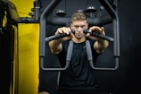 A man is seated at a gym, using a chest press machine. He is wearing a black t-shirt and is focused on his workout. Behind him, the gym equipment is visible against a black background. The lighting highlights the equipment and the man's expression.