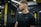 A person with short hair and a beard is at a gym, wearing a black athletic shirt. The environment includes gym equipment and a background with yellow and black color schemes and lighting fixtures.