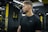 A person with short hair and a beard is at a gym, wearing a black athletic shirt. The environment includes gym equipment and a background with yellow and black color schemes and lighting fixtures.