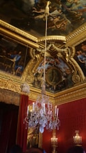An opulent room with a grand chandelier hanging from an intricately decorated ceiling adorned with detailed frescoes and gold embellishments. The walls are lined with rich red curtains and golden ornate carvings.