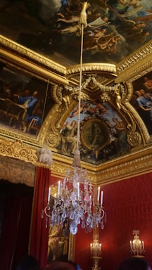 An opulent room with a grand chandelier hanging from an intricately decorated ceiling adorned with detailed frescoes and gold embellishments. The walls are lined with rich red curtains and golden ornate carvings.