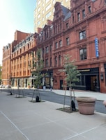 A city street featuring a row of historic, red-brick buildings with intricate architectural details. The buildings have numerous windows and ornate designs. There are small trees planted along the sidewalk and a few pedestrian walkways. The weather appears clear and bright, suggesting a sunny day.