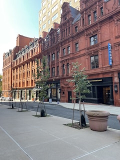 A city street featuring a row of historic, red-brick buildings with intricate architectural details. The buildings have numerous windows and ornate designs. There are small trees planted along the sidewalk and a few pedestrian walkways. The weather appears clear and bright, suggesting a sunny day.
