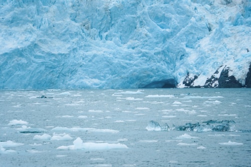 A large, rugged glacier extends across the image, with icy blue surfaces that dominate the landscape. The glacier meets a body of water filled with floating ice chunks. The ice is scattered across the surface, varying in size, creating a cold, arctic environment.