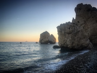Rock formations rise dramatically from the calm sea at sunset, with gentle waves lapping against a rocky shoreline. A yacht is visible on the horizon.