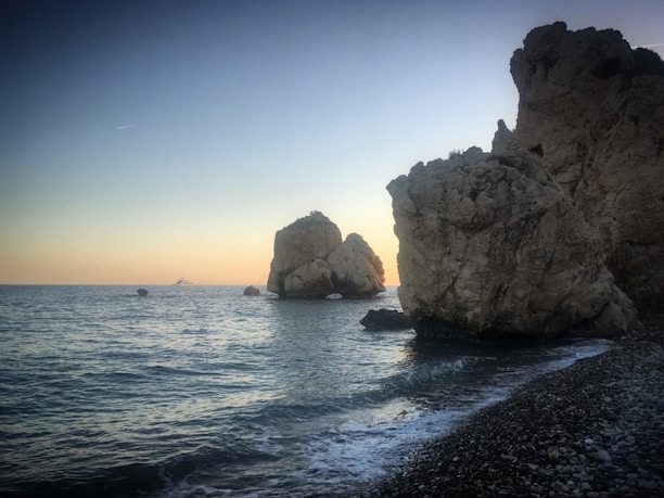 Rock formations rise dramatically from the calm sea at sunset, with gentle waves lapping against a rocky shoreline. A yacht is visible on the horizon.
