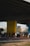 A group of people are gathered on a basketball court under an overpass. The court has a mural featuring a large yellow smiley face with a crown. Players and onlookers are casually dressed, some standing and others seated, creating a laid-back, community atmosphere.