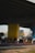 A group of people are gathered on a basketball court under an overpass. The court has a mural featuring a large yellow smiley face with a crown. Players and onlookers are casually dressed, some standing and others seated, creating a laid-back, community atmosphere.