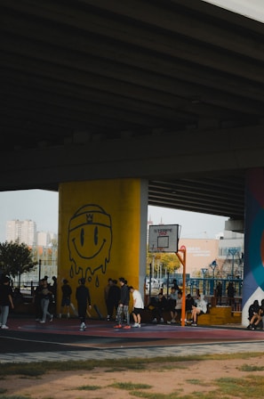 A group of people are gathered on a basketball court under an overpass. The court has a mural featuring a large yellow smiley face with a crown. Players and onlookers are casually dressed, some standing and others seated, creating a laid-back, community atmosphere.