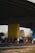 A group of people are gathered on a basketball court under an overpass. The court has a mural featuring a large yellow smiley face with a crown. Players and onlookers are casually dressed, some standing and others seated, creating a laid-back, community atmosphere.