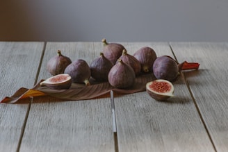 A group of fresh figs is placed on a rustic wooden table, with a tropical leaf underneath. Two figs are cut open, revealing their red, seed-filled interior.