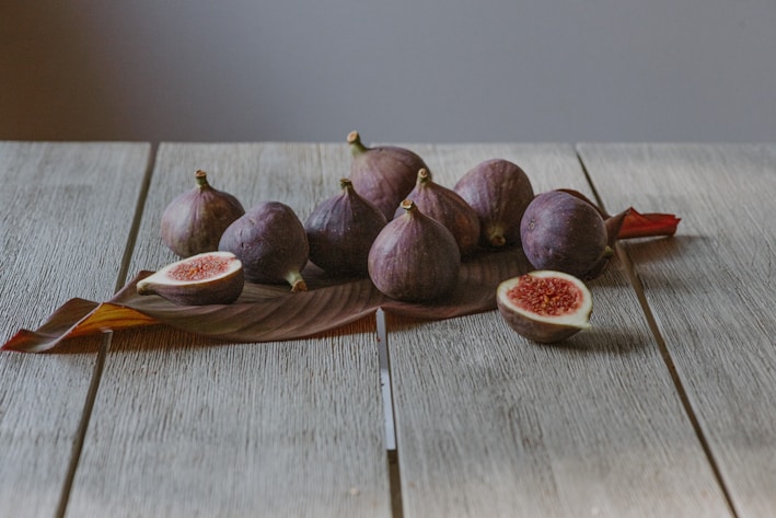 A group of fresh figs is placed on a rustic wooden table, with a tropical leaf underneath. Two figs are cut open, revealing their red, seed-filled interior.