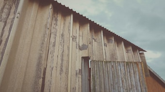 A close-up view of a rustic, weathered metal shed with corrugated iron sheets. The metal has developed significant signs of wear and tear, with streaks of rust and patches of discoloration throughout the surface. The sky in the background is overcast with clouds.