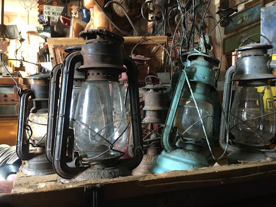 Several rusty, vintage lanterns are arranged on a wooden shelf, surrounded by various antique items in a cluttered setting. The lanterns display signs of age and wear, with some showing a patina of oxidized metal. The background features other nostalgic elements, including old signage and nostalgic decor.