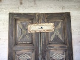A rustic wooden door with intricate geometric carvings, featuring a small sign hung by a rope. The sign is weathered and painted with a minimalist depiction of a sailboat.