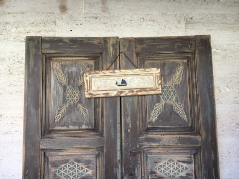 A rustic wooden door with intricate geometric carvings, featuring a small sign hung by a rope. The sign is weathered and painted with a minimalist depiction of a sailboat.