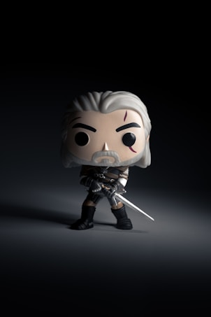 A stylized vinyl figure with white hair and scars, dressed in a dark outfit, holding a sword. The figure stands out against a dark background, casting a shadow on the surface.