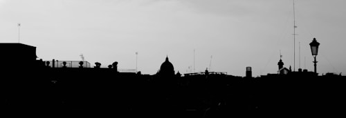 The image depicts a city skyline silhouette against a cloudy sky. Antennas, domes, and varied architectural structures are visible, creating an urban landscape. A street lamp is prominently featured on the right side.