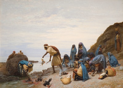 A group of people dressed in blue and brown garments are gathered by the seaside. Some are sitting on the rocky shore while others are standing near the water. Several individuals seem to be engaged in collecting or examining items, possibly fish. There are baskets on the ground filled with items, and a few birds are also visible. The background features a rocky cliff and a cloudy sky.