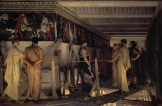 A group of people dressed in ancient attire stands on a wooden platform inside a large, ornately decorated building. The individuals engage in conversation and observation beneath a richly detailed frieze depicting figures in motion. The structure's architecture features intricate patterns along the cornice, and the dim lighting casts soft shadows around the scene.