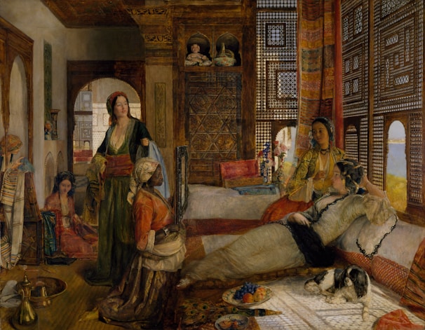 A richly decorated interior featuring several women in traditional attire, relaxed and conversing in an opulent, Eastern-style room. Intricate wooden lattice work and vibrant textiles adorn the room, and a small dog rests peacefully on a carpet. A fruit platter and vases of flowers add to the luxurious atmosphere.