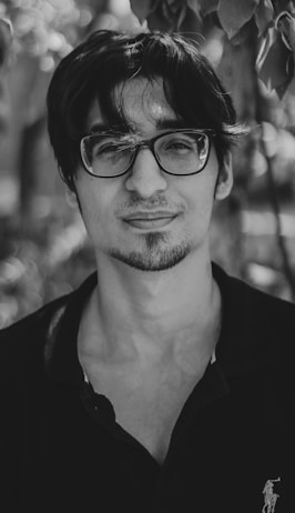 A black and white portrait of a person with dark hair and glasses, wearing a dark shirt. The subject is looking directly at the camera with a neutral expression. The background is blurred with bokeh effect, featuring leaves and natural elements.