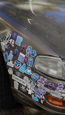 A car's front bumper and side panel are covered with an array of colorful stickers. These include various designs such as cartoon characters, logos, symbols, and abstract art. The car appears to be parked on a paved surface, and the paint shows signs of wear.