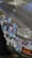A car's front bumper and side panel are covered with an array of colorful stickers. These include various designs such as cartoon characters, logos, symbols, and abstract art. The car appears to be parked on a paved surface, and the paint shows signs of wear.