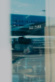 A reflection of an airplane with the word 'PANORAMA' on its side is visible through a large window. Inside, two blurred figures appear to be sitting at a table in a casual setting, suggesting a restaurant or café environment.