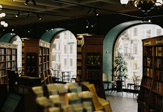 A cozy bookstore interior with tall bookshelves filled with various books. Large arched windows reveal a view of a city street outside. Several chairs and tables are scattered around, inviting visitors to sit and read. Warm lighting creates a welcoming atmosphere, complemented by the presence of a green plant near the window.