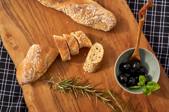 A rustic wooden board with an assortment of olives, bruschetta, and fresh herbs, styled with olive branches on a dark beige table.