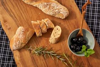 A rustic wooden board with an assortment of olives, bruschetta, and fresh herbs, styled with olive branches on a dark beige table.
