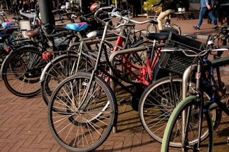 A group of bicycles tightly parked together on a brick-paved area. Some bikes have accessories like baskets and child seats. In the background, a few people can be seen walking, and one person is riding a bicycle.