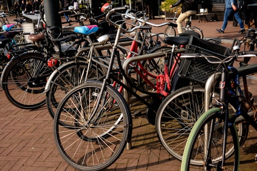 A group of bicycles tightly parked together on a brick-paved area. Some bikes have accessories like baskets and child seats. In the background, a few people can be seen walking, and one person is riding a bicycle.