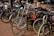 A group of bicycles tightly parked together on a brick-paved area. Some bikes have accessories like baskets and child seats. In the background, a few people can be seen walking, and one person is riding a bicycle.
