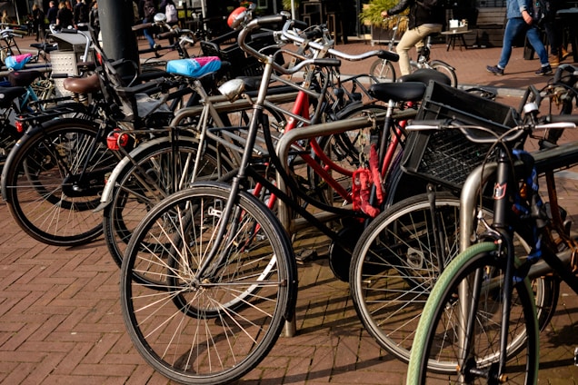 A group of bicycles tightly parked together on a brick-paved area. Some bikes have accessories like baskets and child seats. In the background, a few people can be seen walking, and one person is riding a bicycle.