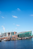 A modern green building with a sloped roof resembling a ship's hull, set along a waterfront with several docked boats. The sky is bright blue with a few scattered clouds. In the background, there are multi-story contemporary buildings.