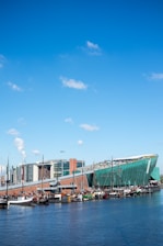 A modern green building with a sloped roof resembling a ship's hull, set along a waterfront with several docked boats. The sky is bright blue with a few scattered clouds. In the background, there are multi-story contemporary buildings.