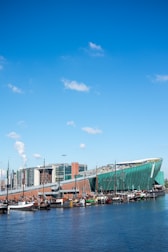 A modern green building with a sloped roof resembling a ship's hull, set along a waterfront with several docked boats. The sky is bright blue with a few scattered clouds. In the background, there are multi-story contemporary buildings.