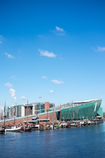 A modern green building with a sloped roof resembling a ship's hull, set along a waterfront with several docked boats. The sky is bright blue with a few scattered clouds. In the background, there are multi-story contemporary buildings.