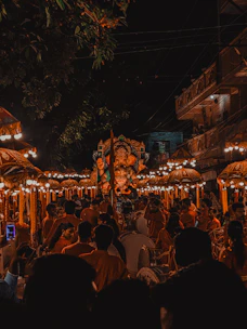 A large crowd gathers at night for a vibrant cultural celebration featuring a prominent statue of a deity adorned with lights and decorations. The scene is illuminated by numerous light fixtures, creating a festive atmosphere. People dressed in bright clothing, some playing musical instruments, are seen participating in the procession. Ornate umbrellas and trees frame the scene.