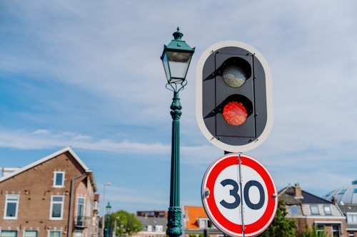 A traffic light with a red signal is positioned next to a speed limit sign indicating 30. The background includes a blue sky and residential buildings with pitched roofs.