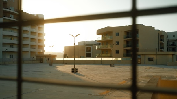 Urban residential buildings seen through a wire fence during sunset, casting long shadows on a concrete surface. The architecture is modern with multiple balconies and the area appears quiet and deserted.