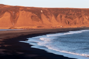 A dramatic coastal landscape with black sand beaches meeting the vibrant blue ocean. In the background, there are towering orange-brown cliffs bathed in warm sunlight, creating a stark contrast with the dark shoreline. White waves gently crash along the shore, adding an element of motion to the serene setting. Sparse structures can be seen at the base of the cliffs.