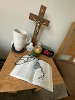 A small wooden table holds a variety of personal items including an open book, likely a Bible, with a rosary and a pair of glasses resting on it. Other items on the table include a mug with the words 'pray more worry less,' a small figurine or container, a green candle in a glass jar, a digital clock showing 3:00, and a decorative stone with the word 'Hope' inscribed. Above the table, mounted on the wall, is a crucifix.