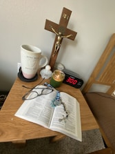 A small wooden table holds a variety of personal items including an open book, likely a Bible, with a rosary and a pair of glasses resting on it. Other items on the table include a mug with the words 'pray more worry less,' a small figurine or container, a green candle in a glass jar, a digital clock showing 3:00, and a decorative stone with the word 'Hope' inscribed. Above the table, mounted on the wall, is a crucifix.
