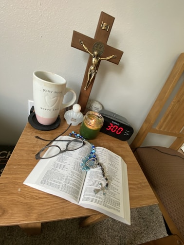 A small wooden table holds a variety of personal items including an open book, likely a Bible, with a rosary and a pair of glasses resting on it. Other items on the table include a mug with the words 'pray more worry less,' a small figurine or container, a green candle in a glass jar, a digital clock showing 3:00, and a decorative stone with the word 'Hope' inscribed. Above the table, mounted on the wall, is a crucifix.